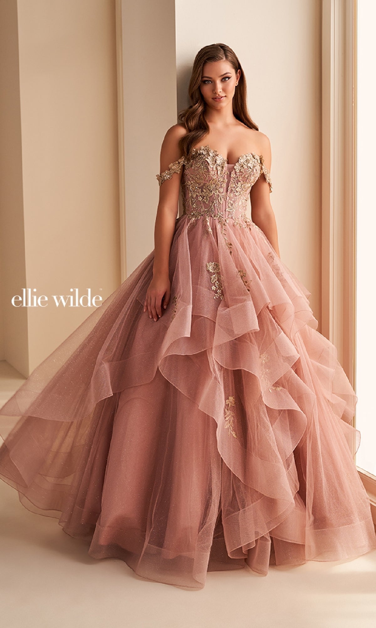 A woman stands by a window wearing the Ellie Wilde Long Designer Prom Ball Gown EW35084 in blush pink, with floral embroidery and a tiered glitter tulle skirt. The